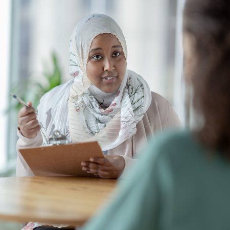 A female Muslim Therapist sits across a desk from her young teenage patient as they talk about her concerns and struggles.  She is dressed casually and wearing a Hijab as she holds a clipboard and takes notes from the meeting.  The teen is dressed casually in a green t-shirt and sitting across from the therapist.